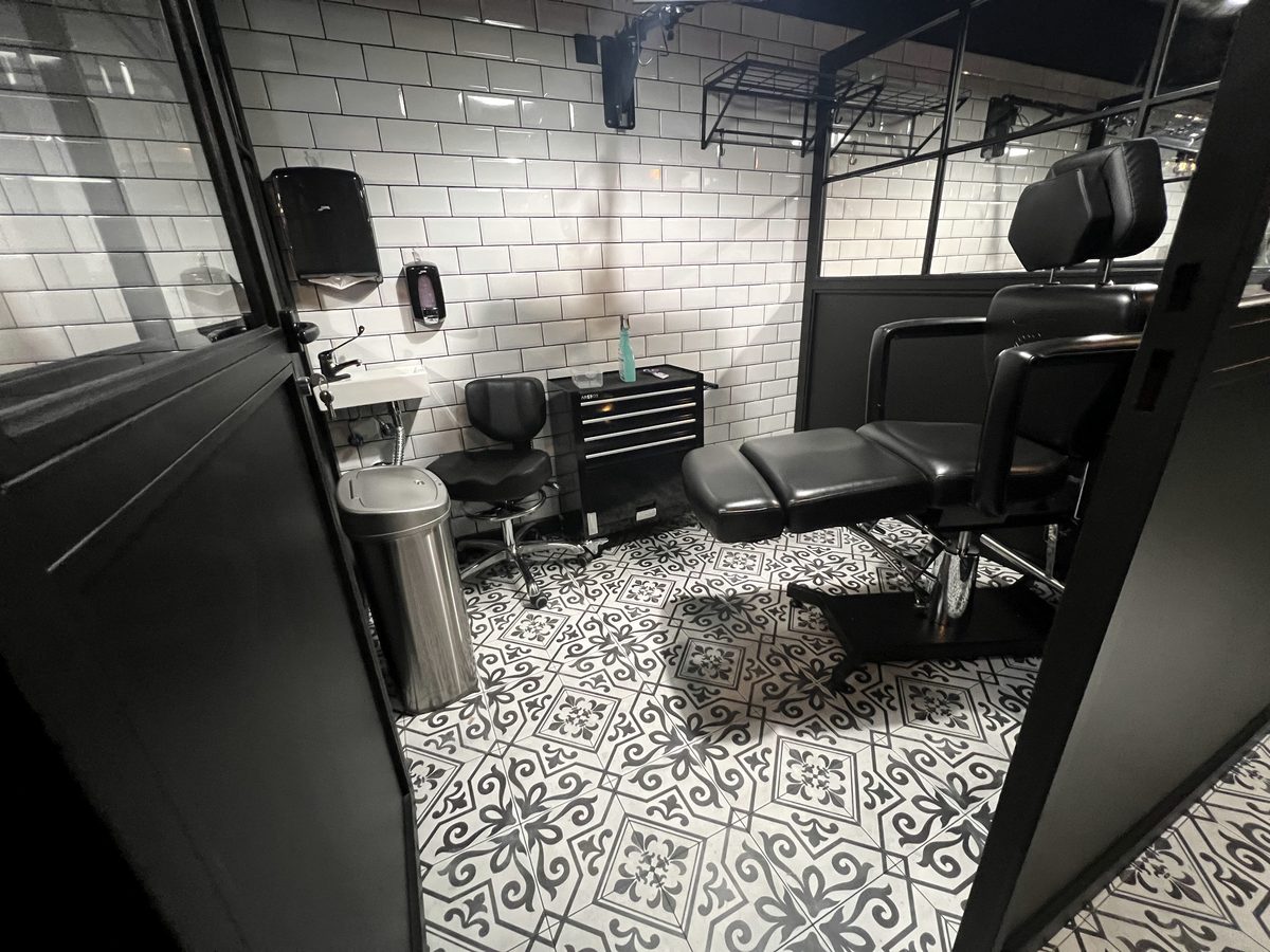 Cabina de tatuaje de Calipso alicatada en azulejo blanco, con camilla negra, lavabo y carro de material.